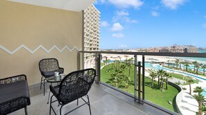Ein Balkon mit zwei Korbstühlen, einem kleinen Tisch und Blick auf ein Resort mit Pool und Palmen.