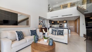 Un salon moderne avec un canapé, une table basse et des objets de décoration. On aperçoit une cuisine à l’arrière-plan, équipée d’appareils électroménagers modernes.