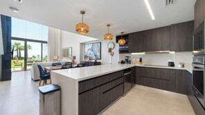 Eine moderne Küche mit dunklen Holzsäcken, einer weißen Arbeitsplatte und einer zentralen Kochinsel. Es gibt einen Essbereich mit Tisch und Stühlen sowie ein großes Fenster mit Blick auf Palmen.