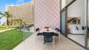 A modern hotel with a curved exterior, a dining area with a white table and chairs, and a view of palm trees and a building in the background. - One 101 : 👑 Azure Royale — upscale and memorable (Puerto Peñasco)