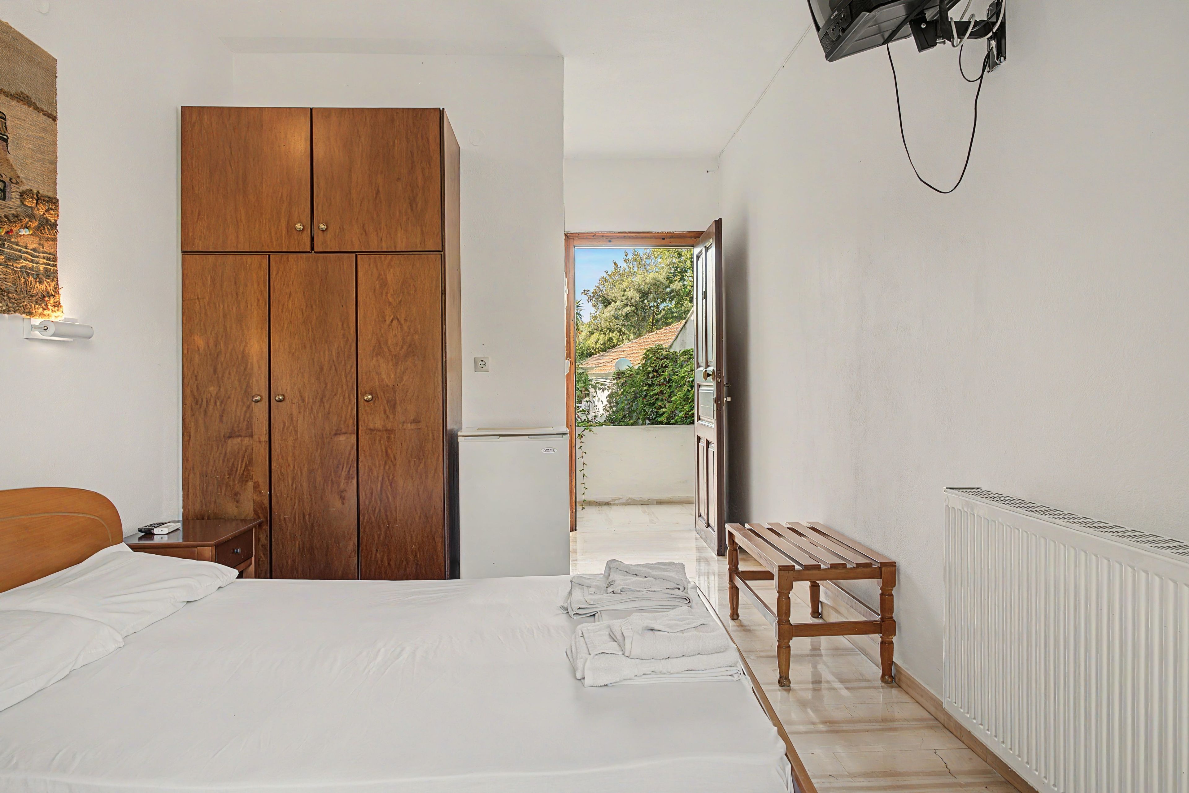 Quarto com cama, guarda-roupa de madeira, um banquinho e vista para o exterior.