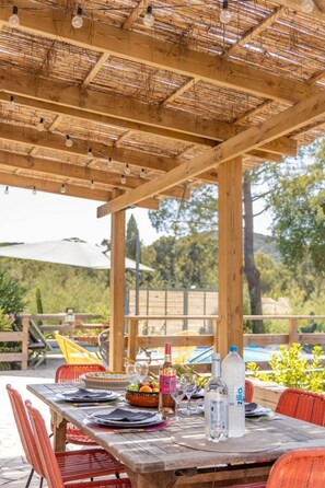 Una pérgola de madera al aire libre con luces colgantes, una mesa lista para comer con sillas y vistas a la vegetación de fondo.