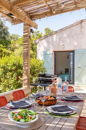 An outdoor dining area with a table set for two, a grill, and a view of a house with shutters. - Villa Gioia - SAN GAVINO DI CARBINI (SAN GAVINO DI CARBINI)
