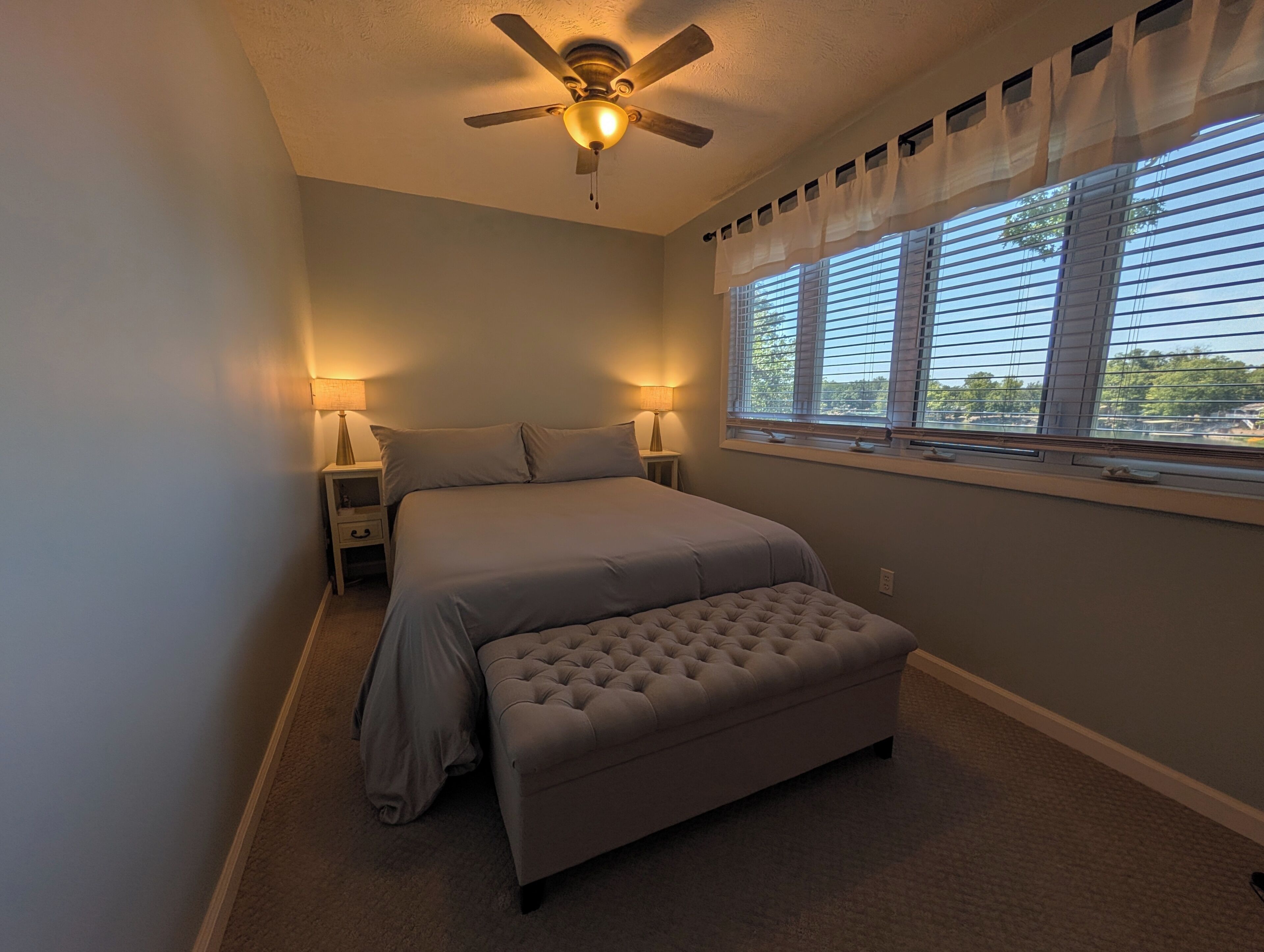 A bedroom with a bed, bedside lamps, a ceiling fan, a window with blinds, and a carpeted floor.