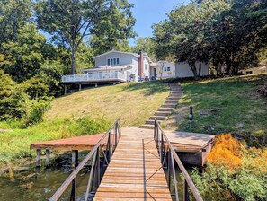 A wooden dock leading to a house with a deck, surrounded by greenery and a small water body.