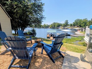 A lakeside patio with Adirondack chairs, a fire pit, and a view of a dock and boat.