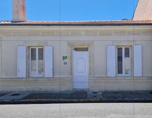 Un edificio con puerta blanca, dos ventanas con persianas y un cartel pequeño en la puerta.