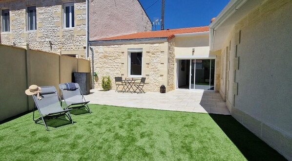 A courtyard with artificial grass, two lounge chairs, a small table, and a door leading to a covered area. - Vrbo Property (LESPARRE MEDOC)