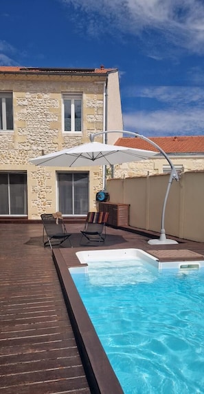 Una piscina con muelle de madera, dos sillas de descanso y una sombrilla blanca.
