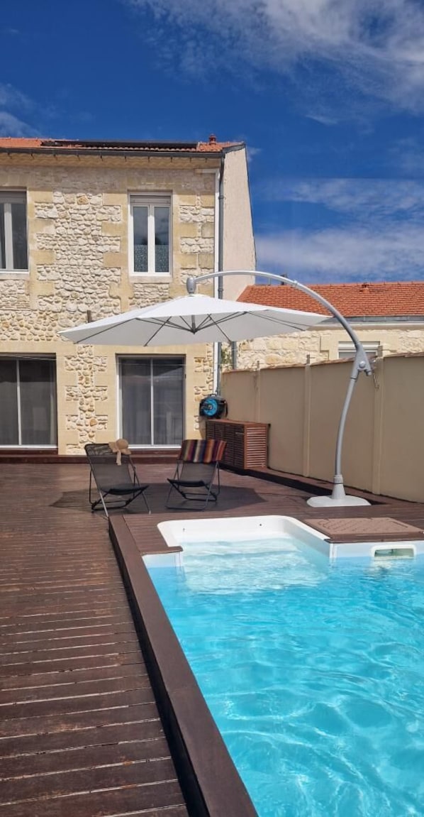 Piscina com deck de madeira, duas espreguiçadeiras e um guarda-sol branco.