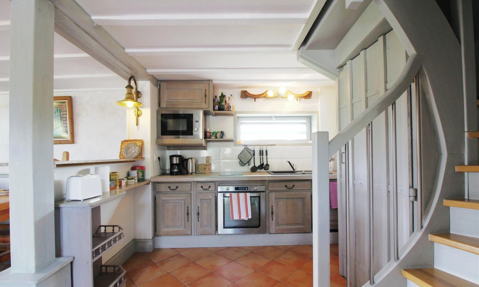 Une cuisine équipée d’un four à micro-ondes, d’un four traditionnel et de divers appareils électroménagers. Il y a une fenêtre au-dessus de l’évier et un escalier sur la droite.