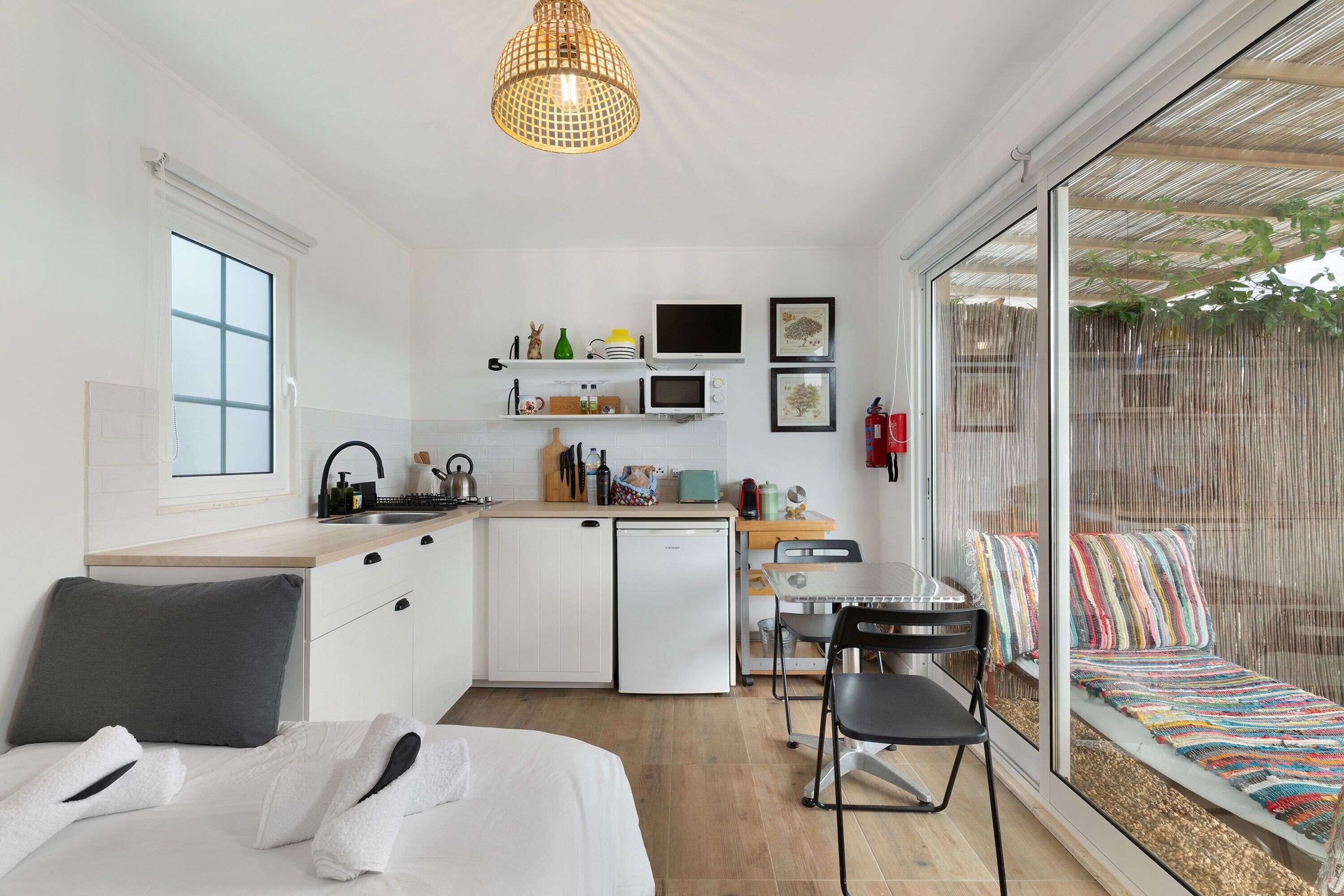A compact kitchen with a sink, microwave, and small refrigerator. There is a dining area with a table and chairs, and a large window with a view of a garden.