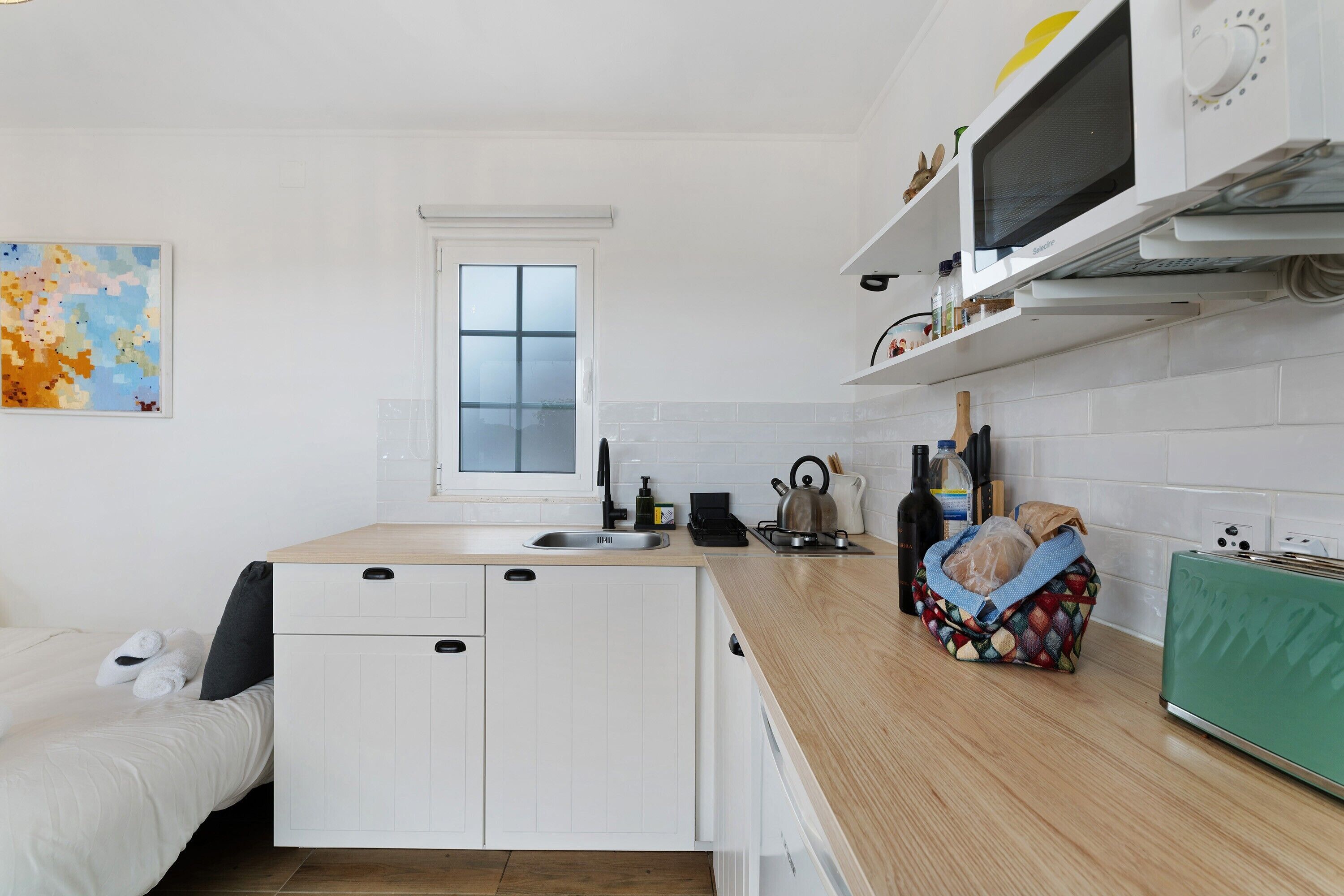 A compact kitchen with a window, white cabinets, a microwave, and a kettle on the counter.