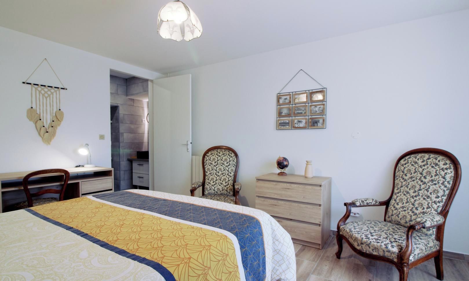 Quarto com cama, duas cadeiras, cômoda, escrivaninha e luminária na parede.