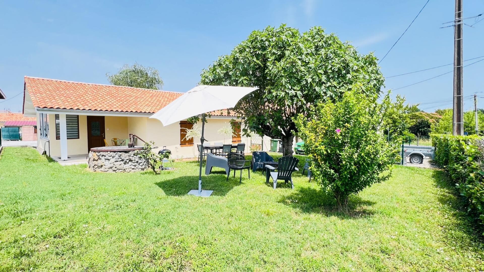 Une maison avec un toit en tuiles, une terrasse avec des meubles d’extérieur et un arbre qui offre de l’ombre.