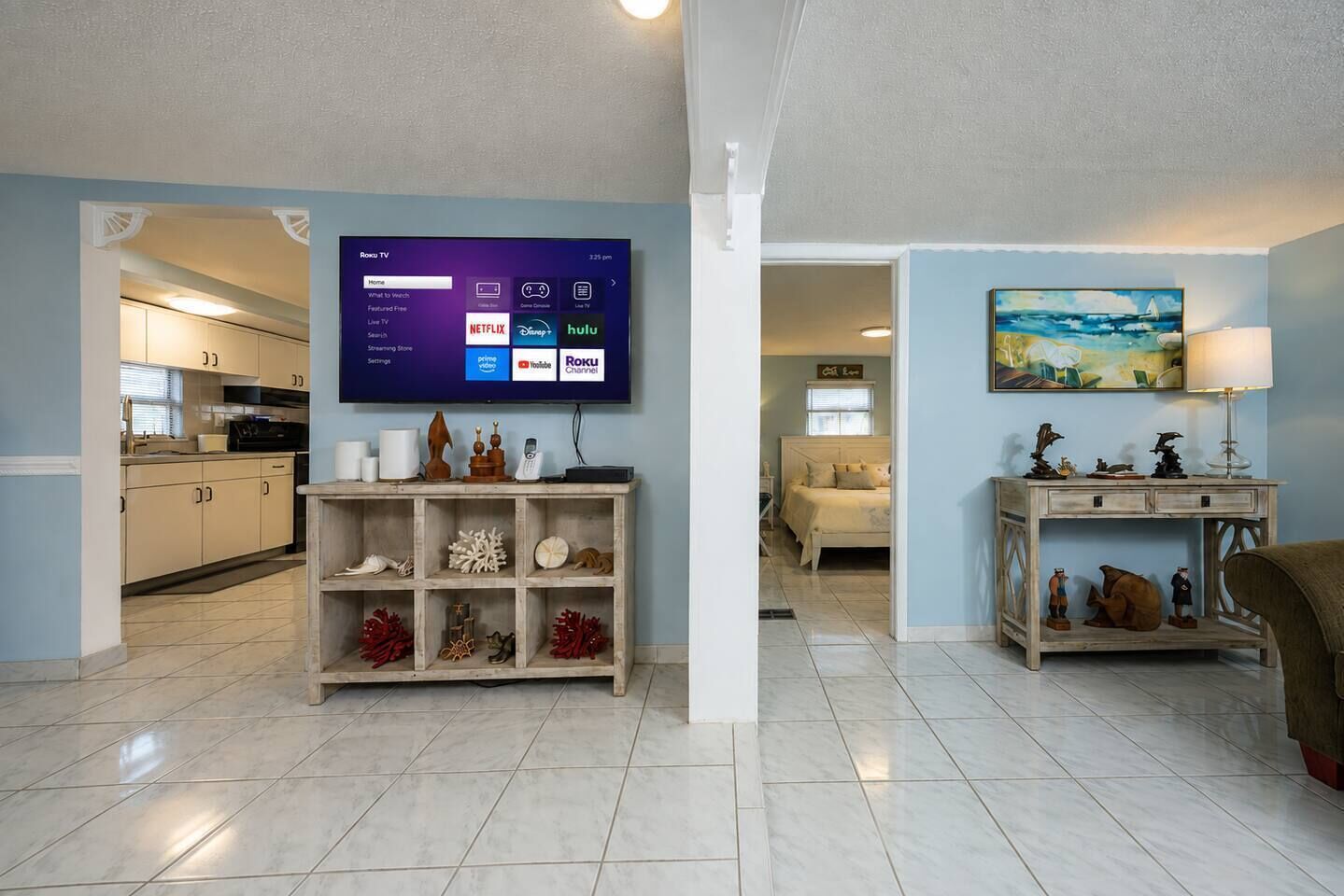 Una sala moderna con un televisor de pantalla plana montado en la pared, una mesa consola de madera con objetos decorativos, una cama con cabecera y un cuadro en la pared.
