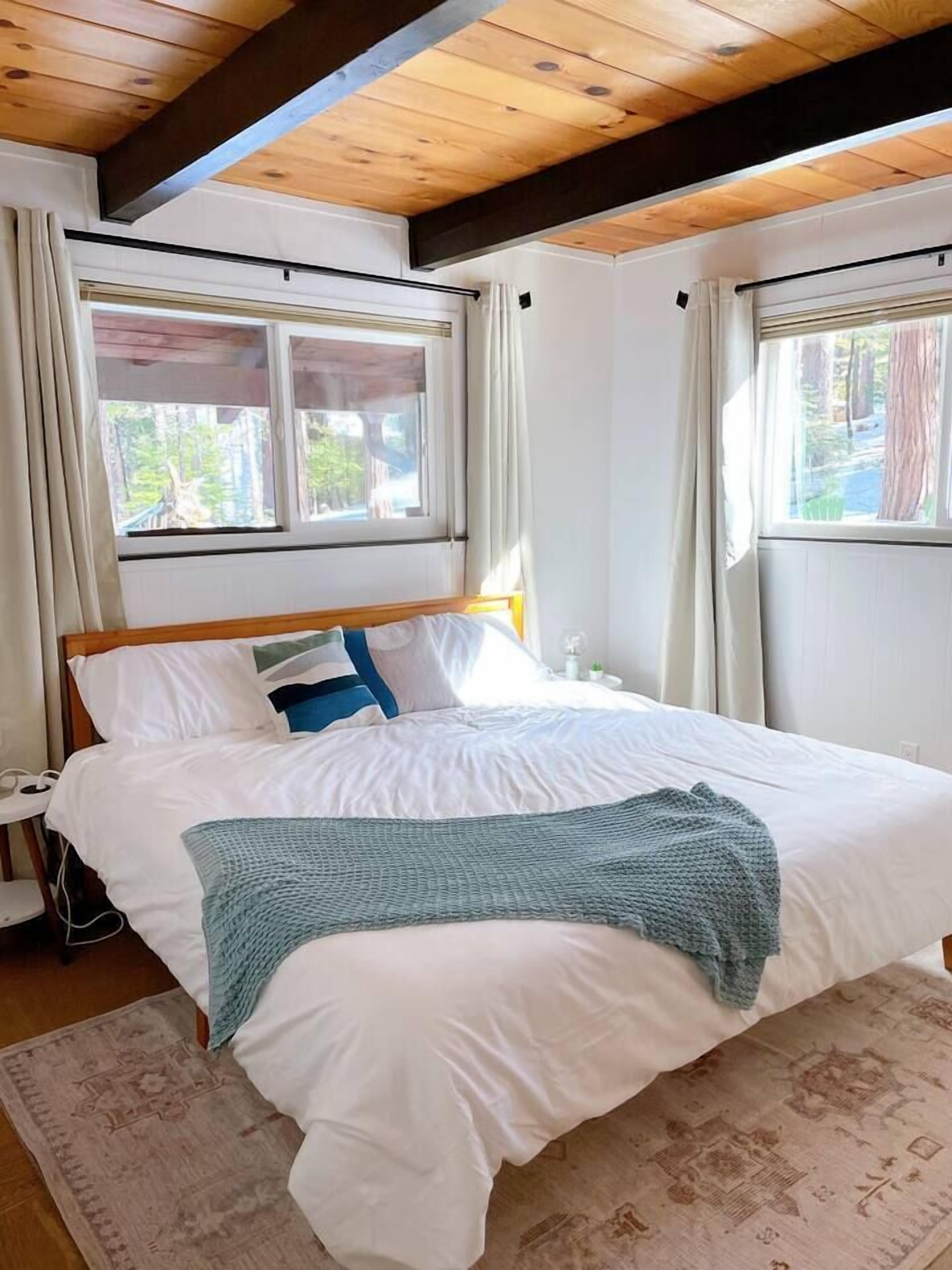 A bedroom with a wooden ceiling, a bed with white bedding, a blue knitted throw, and a patterned rug.