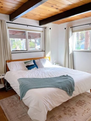 Un dormitorio con techo de madera, cama con ropa de cama blanca, una manta azul de punto y una alfombra estampada.