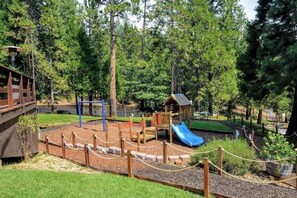 A playground with a slide, climbing structures, and a small building, surrounded by trees.