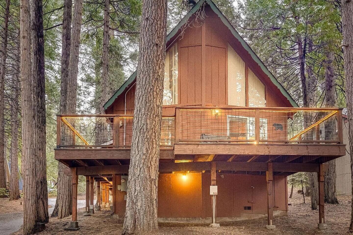 Een houten chalet met een balkon en een benedenverdieping, omgeven door hoge bomen.
