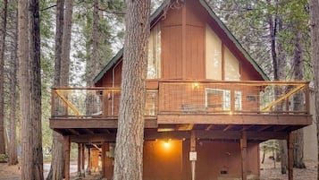 Een houten chalet met een balkon en een benedenverdieping, omgeven door hoge bomen.