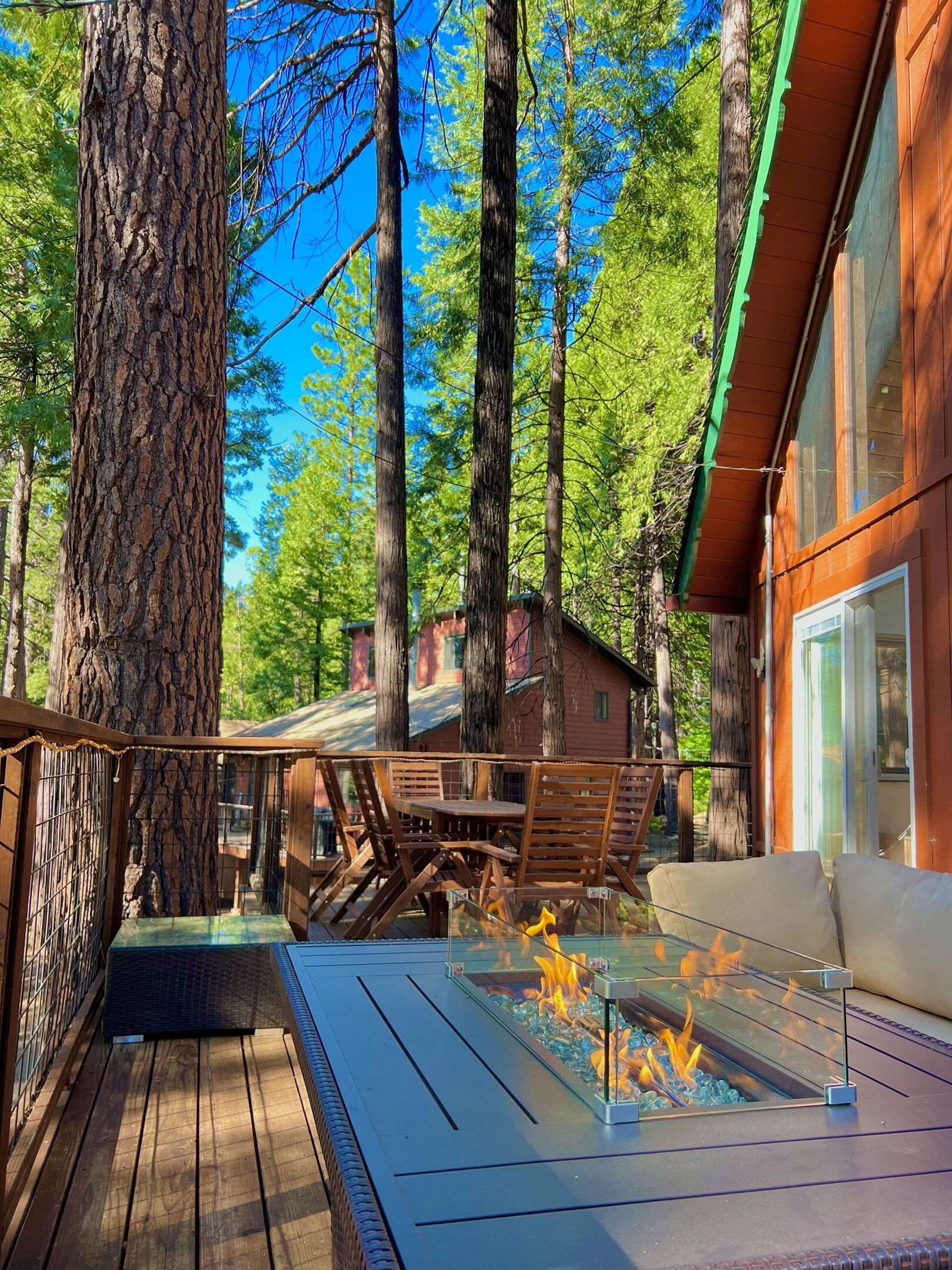 A deck with a glass-top fire pit, wooden furniture, and a view of a forested area.