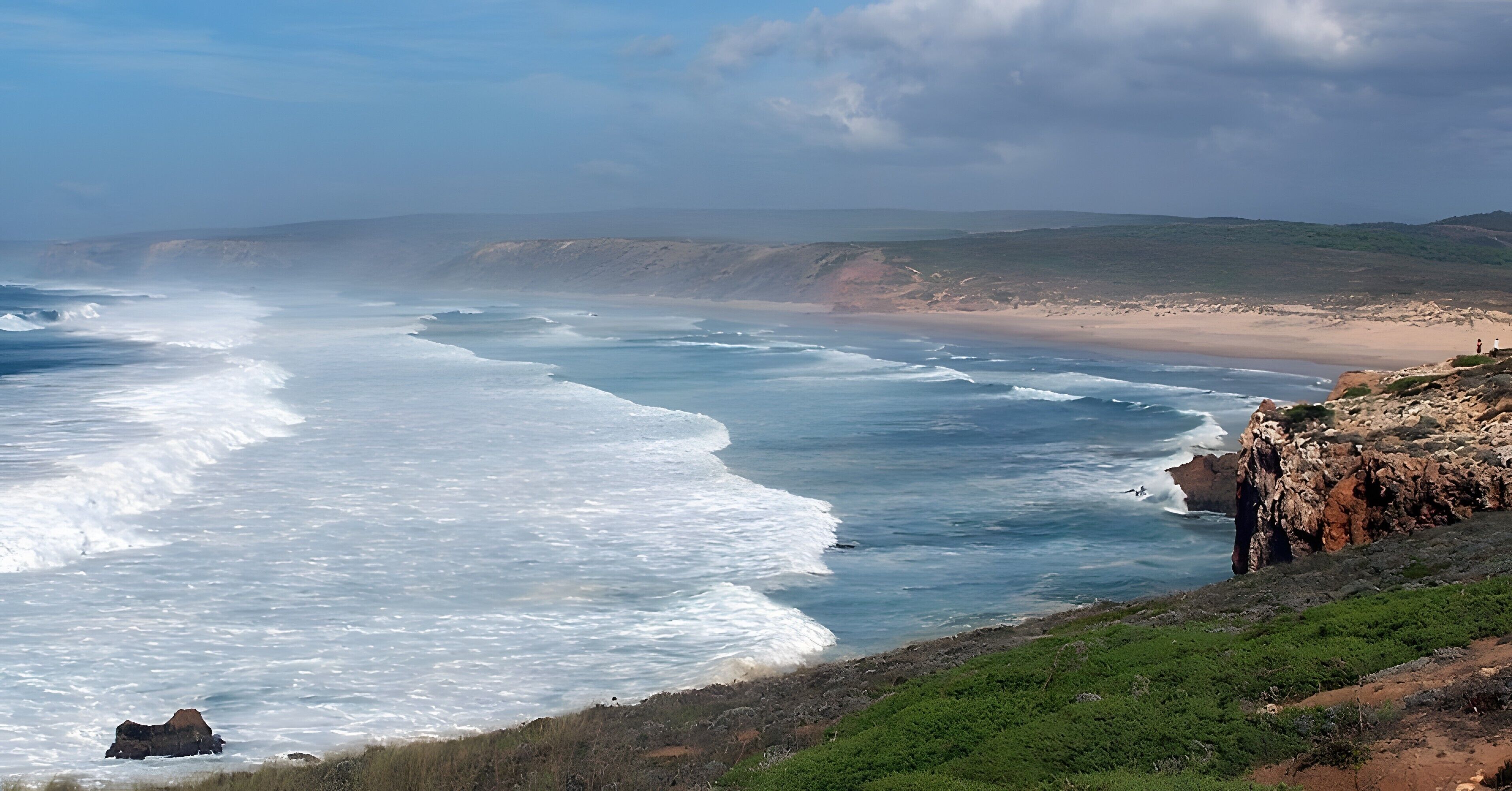 A coastal landscape with waves crashing onto a rocky shore, a sandy beach, and a hilly terrain in the background.