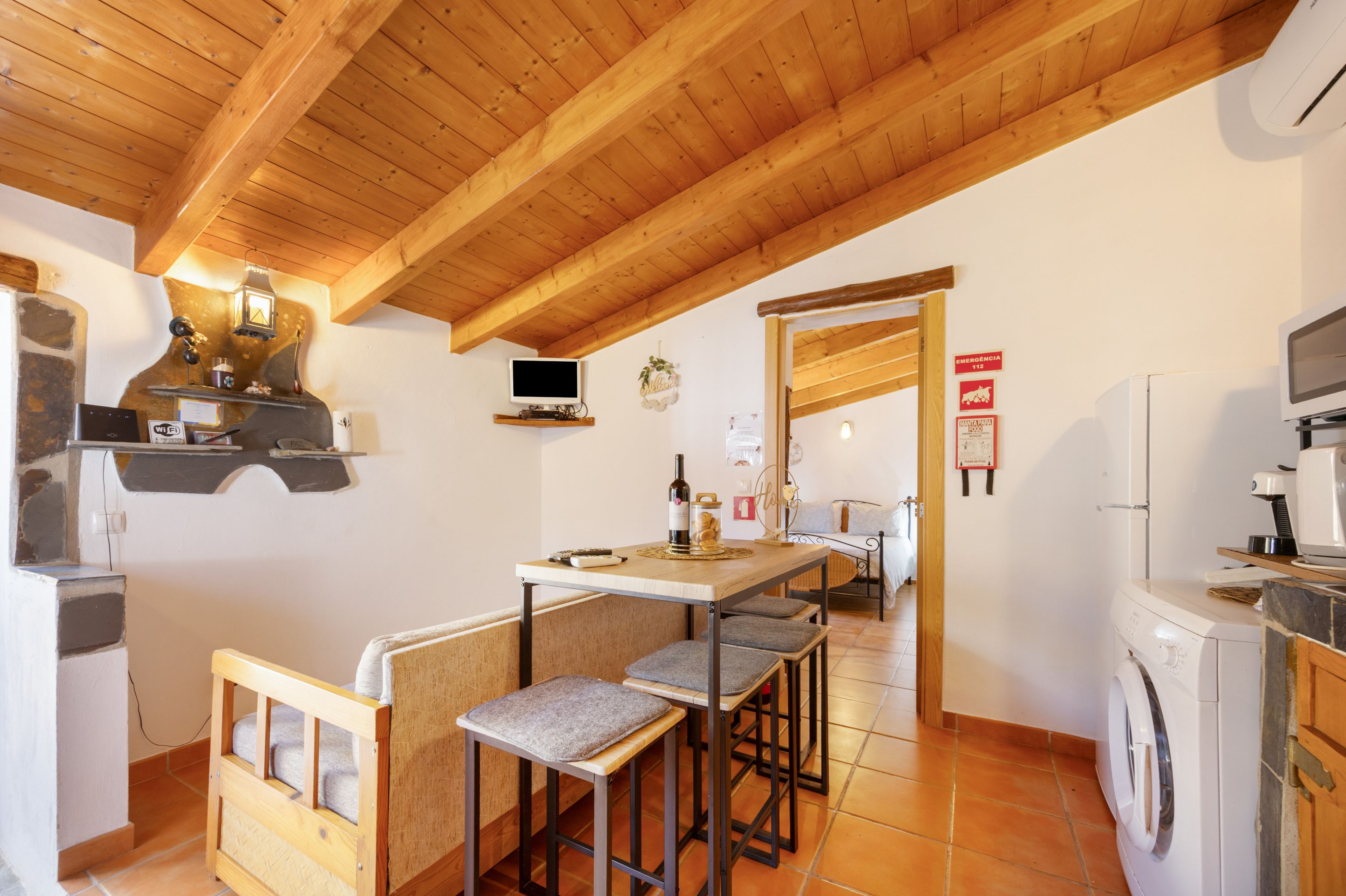 A compact kitchen with a wooden ceiling, a dining area with a table and chairs, a washing machine, and a television on the wall.