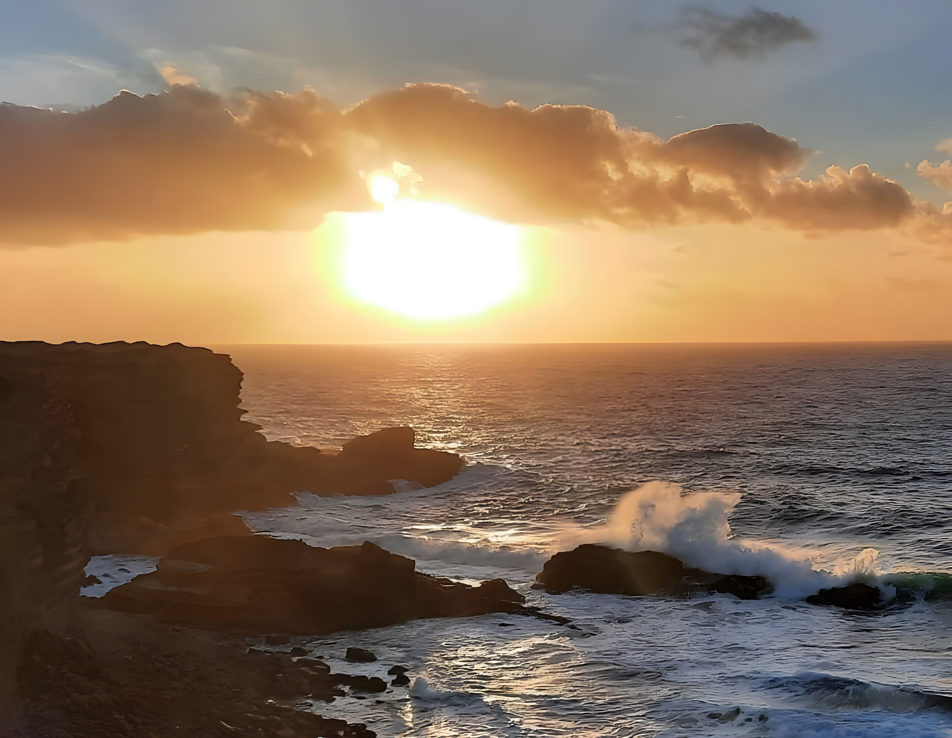 Atardecer costero con el sol bajo en el horizonte, olas rompiendo contra las rocas y un cielo despejado.