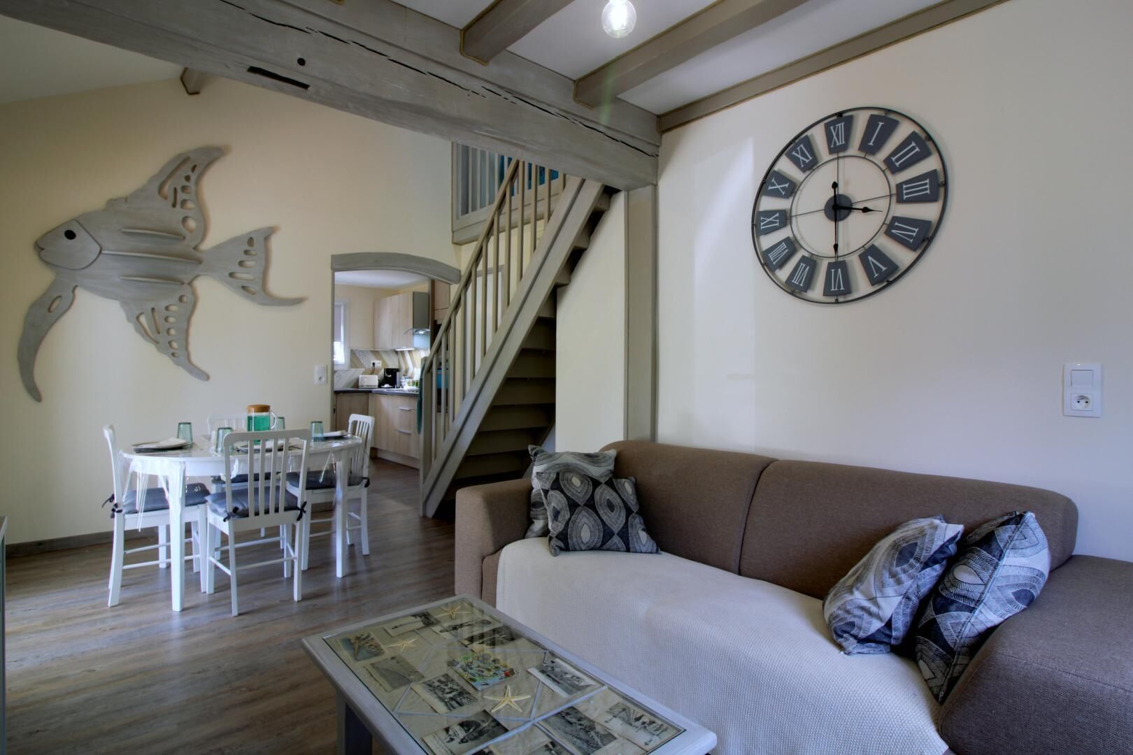 Una sala de estar con un sofá marrón, un reloj de pared redondo, un comedor con mesa y sillas blancas, y una escalera que conduce a otra habitación.