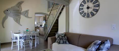 Ein Wohnzimmer mit einem braunen Sofa, einer runden Wanduhr, einem Essbereich mit weißem Tisch und Stühlen sowie einer Treppe zu einem anderen Raum.