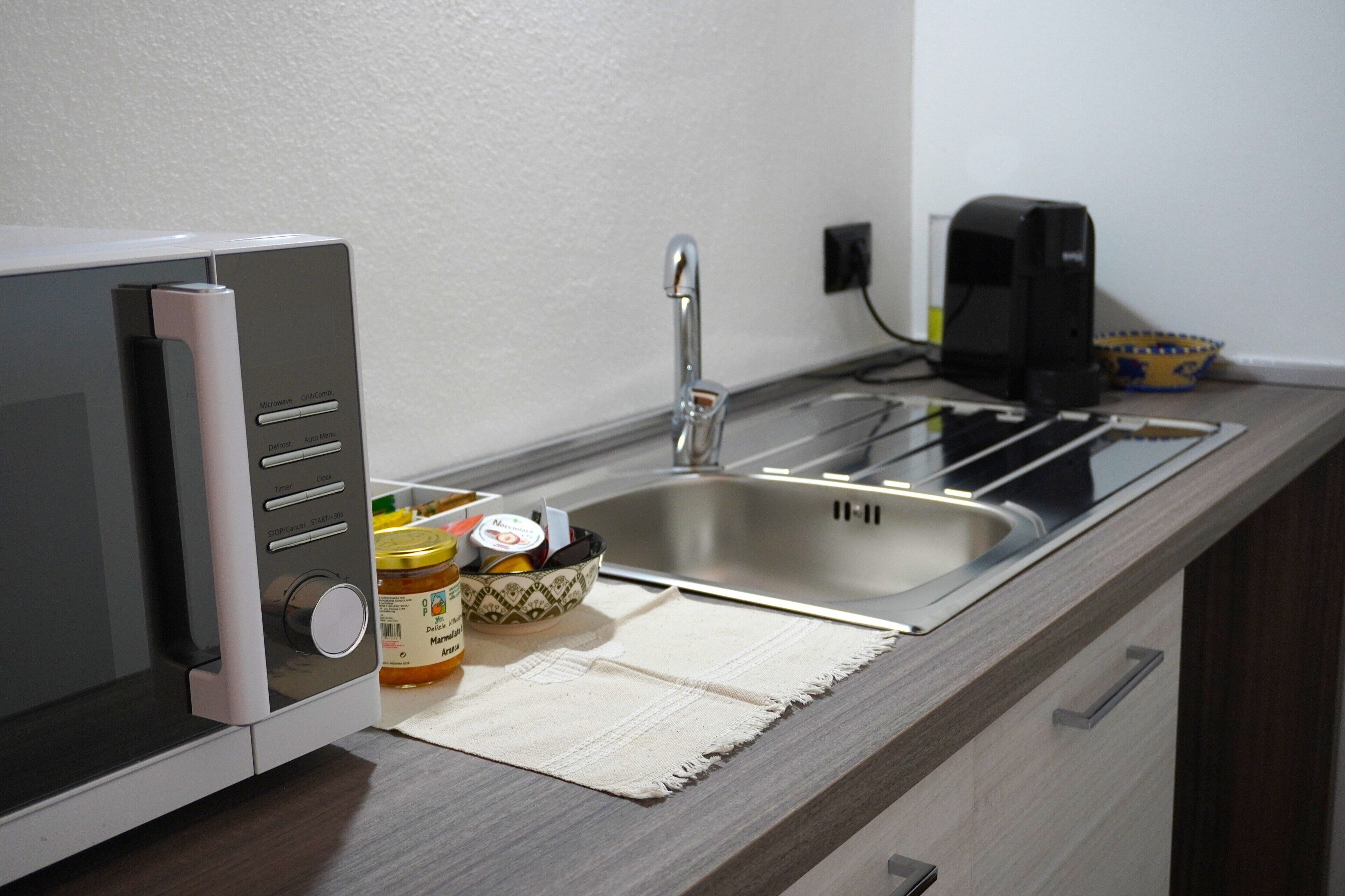 A kitchen with a microwave, a sink, and a coffee maker. There are jars of jam and a bottle of water on the counter.