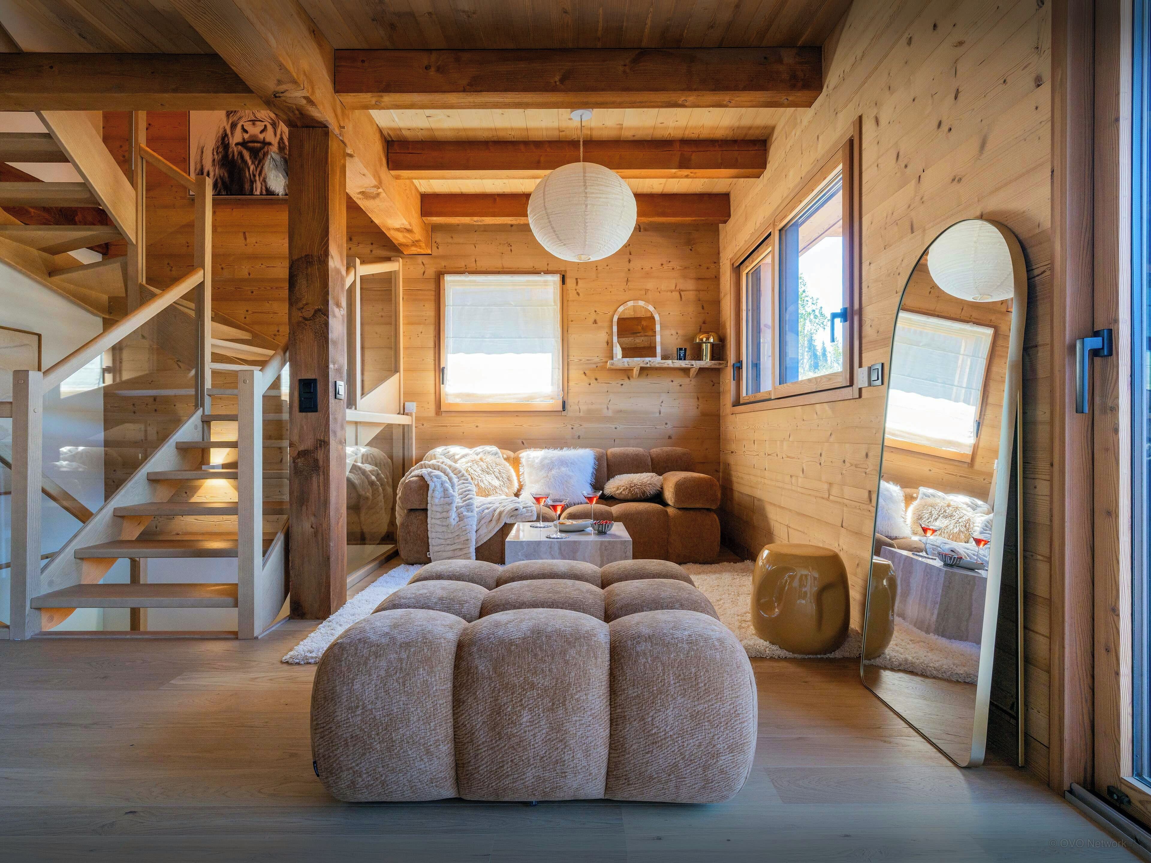 Un intérieur de chalet chaleureux avec un escalier en bois, un coin salon confortable et de grandes fenêtres.