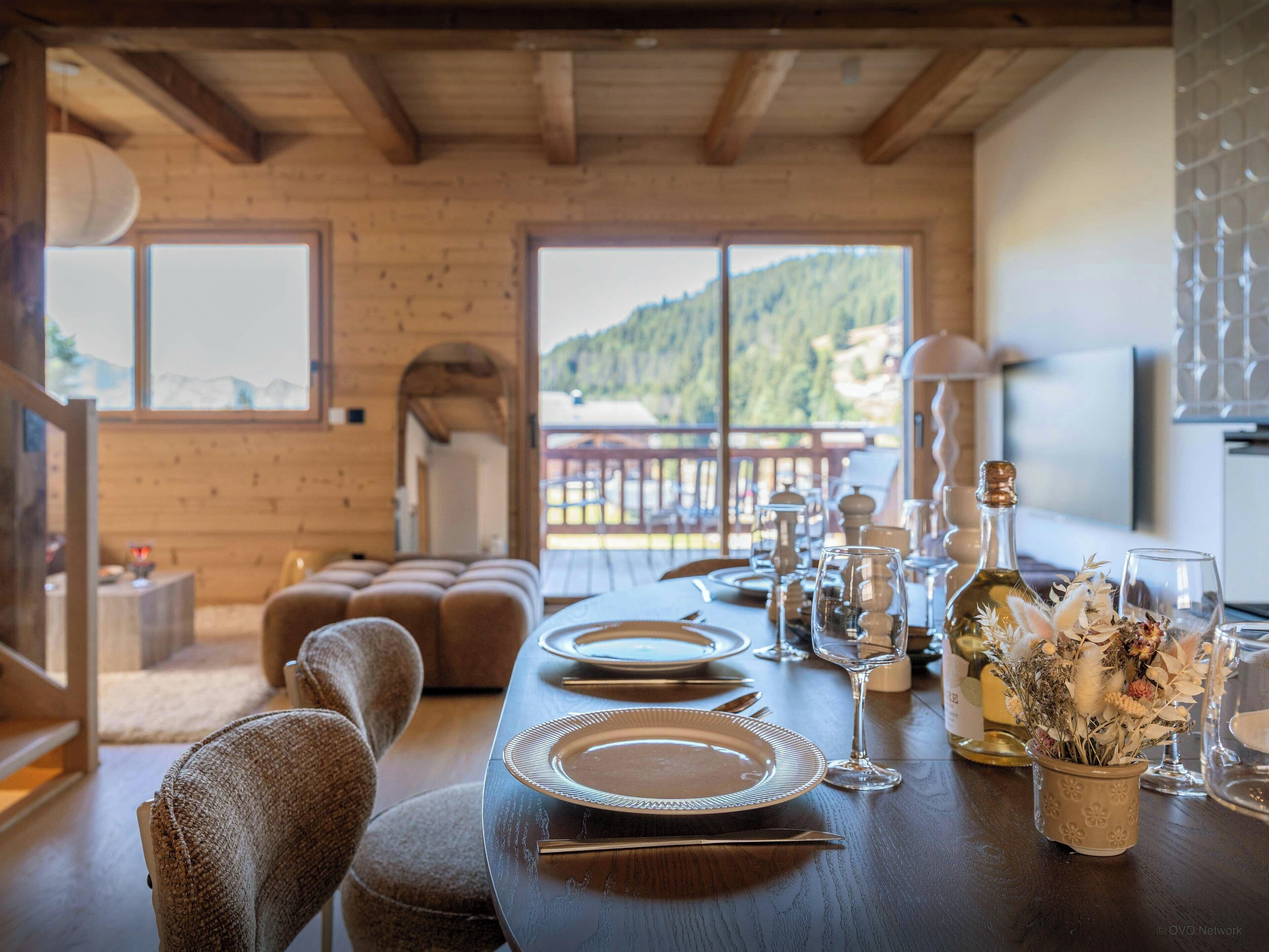 Un comedor con mesa de madera dispuesta para una comida, un sofá y vistas a un paisaje montañoso a través de amplios ventanales.