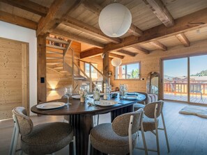 Un intérieur en bois avec une table à manger dressée pour quatre personnes, entourée de chaises en osier, d’un escalier et d’une grande fenêtre donnant sur l’extérieur.