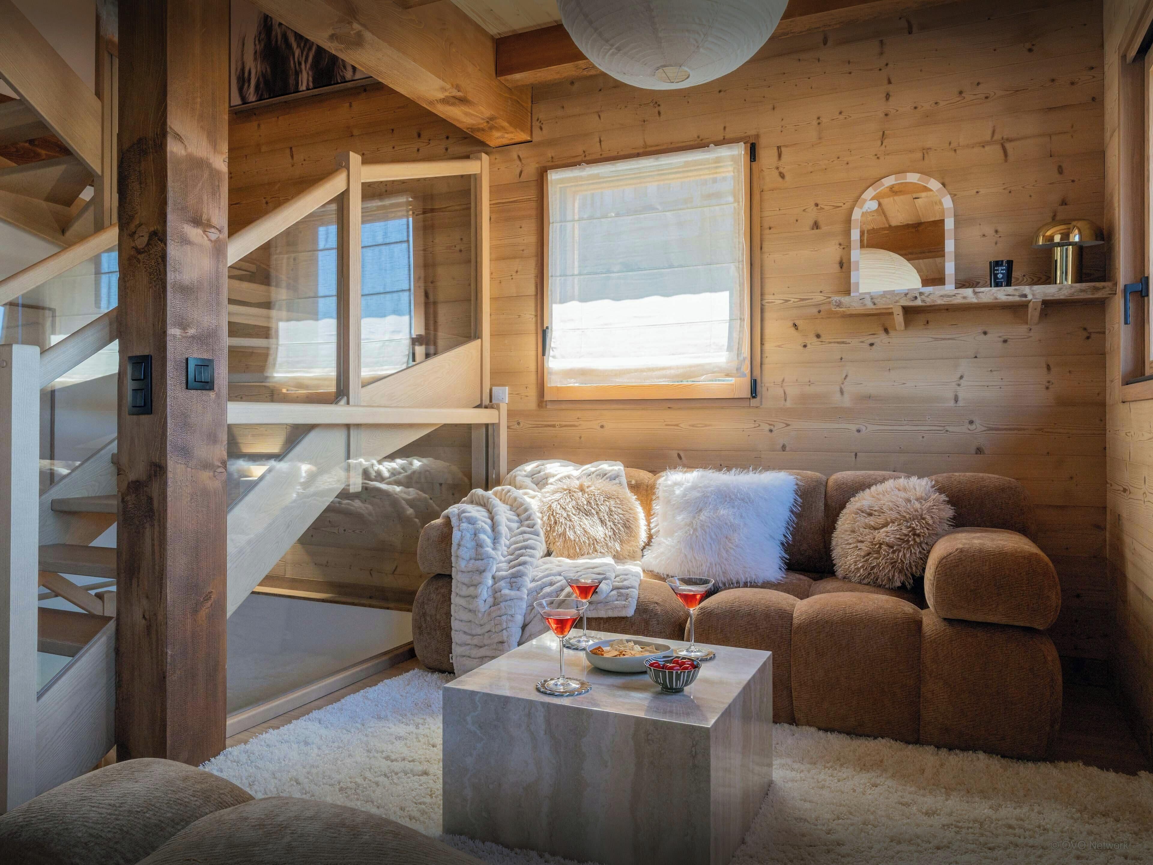 L’intérieur chaleureux d’un chalet avec un escalier en bois, un coin salon confortable avec des coussins et une table basse en marbre où sont disposés des en-cas et des boissons.