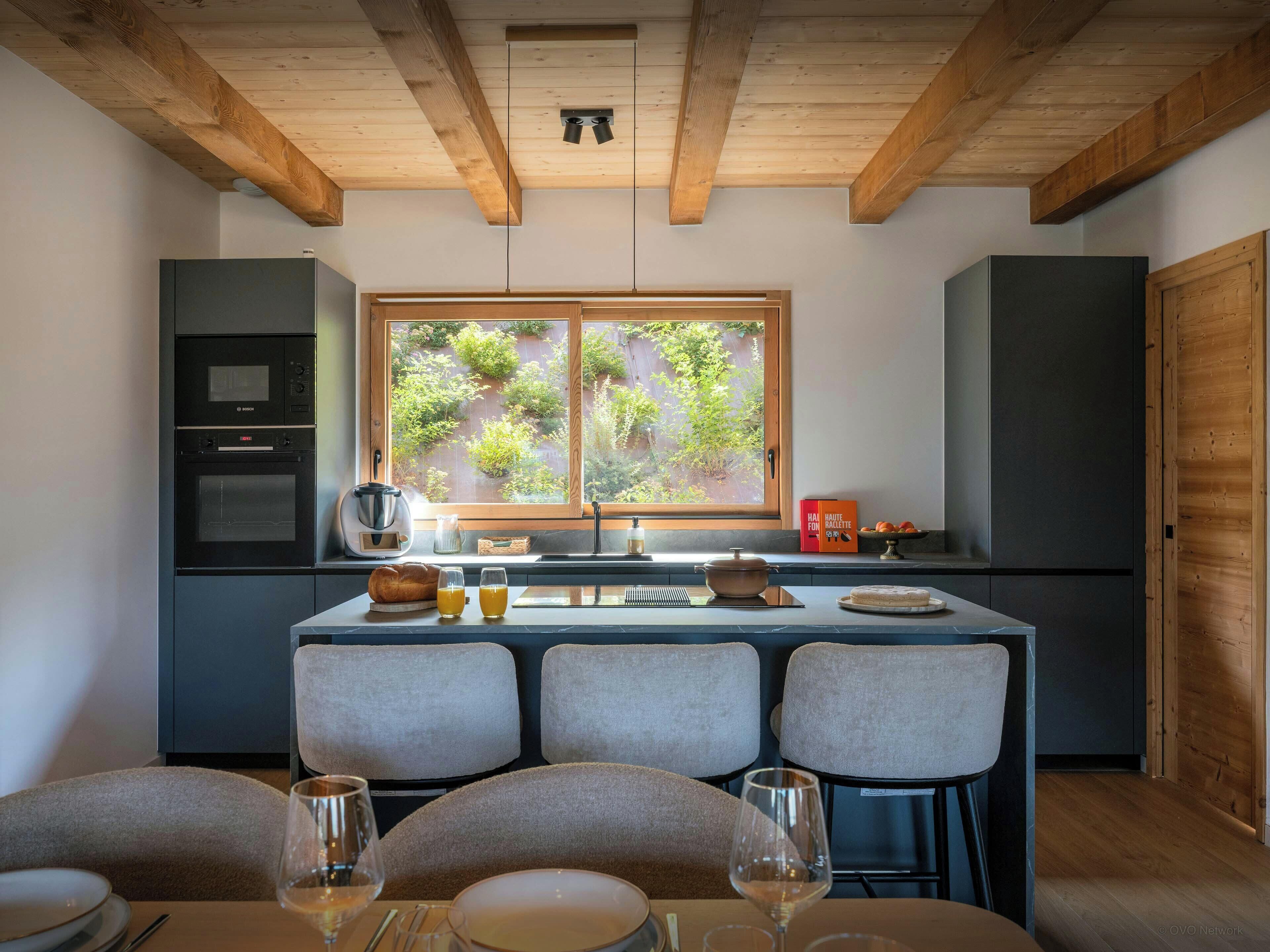 Una cocina moderna con comedor, destacando un techo de madera, un ventanal con vistas a vegetación y un horno empotrado.