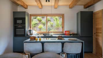 Una cocina moderna con comedor, destacando un techo de madera, un ventanal con vistas a vegetación y un horno empotrado.