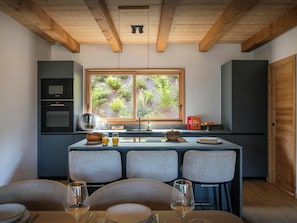 A modern kitchen with a dining area, featuring a wooden ceiling, a large window with a view of greenery, and a built-in oven.