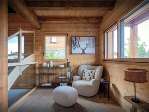 A cozy cabin interior with a wooden interior, a comfortable chair, a round ottoman, a small table, and a window with a view of the outside.