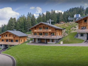 A wooden chalet with a balcony, set against a backdrop of trees and a clear sky.