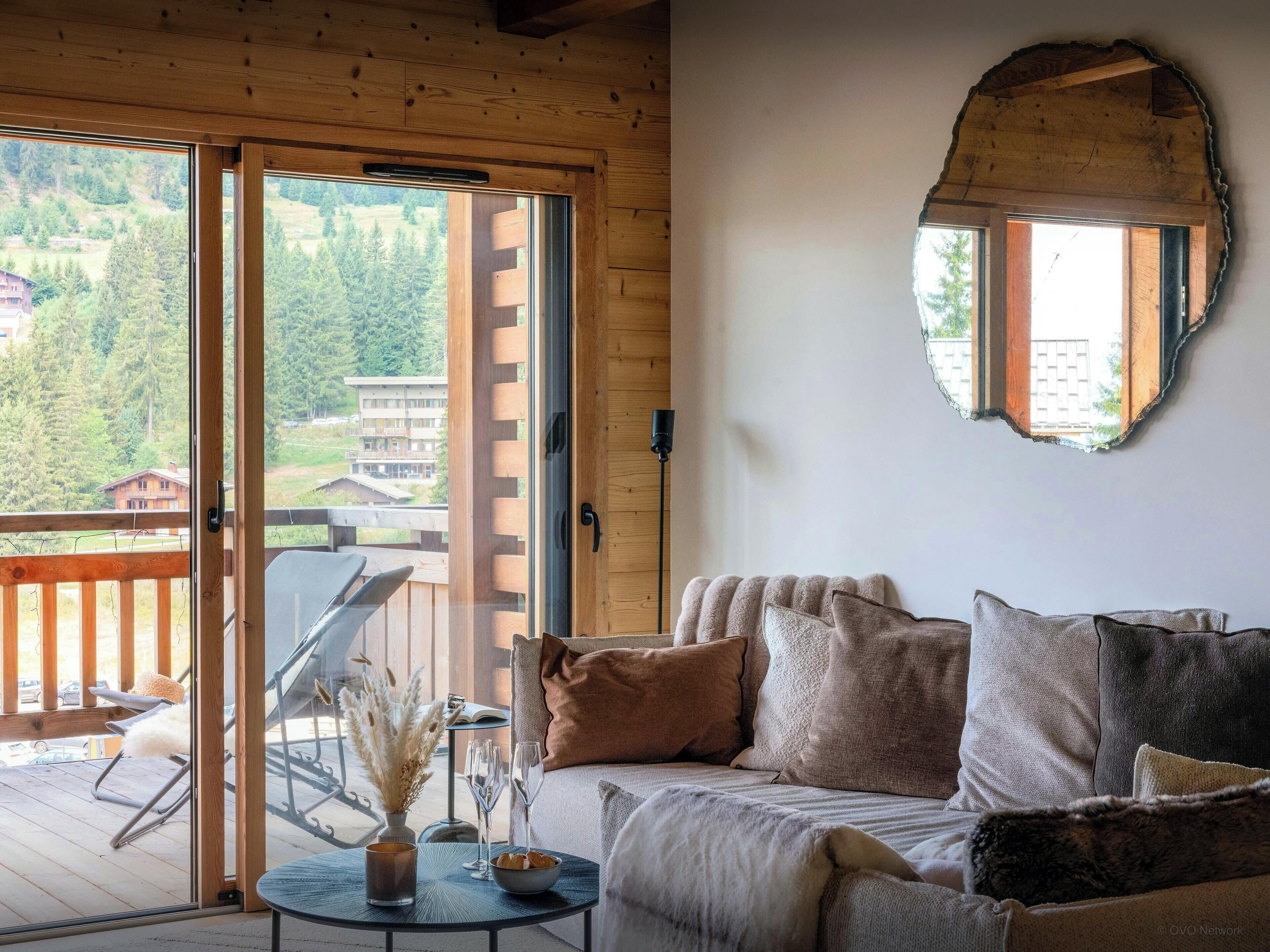 Une pièce chaleureuse avec un plafond en bois, une porte-fenêtre coulissante donnant sur un balcon, un canapé avec des coussins, une petite table avec une bougie et des verres, et un miroir décoratif accroché au mur.