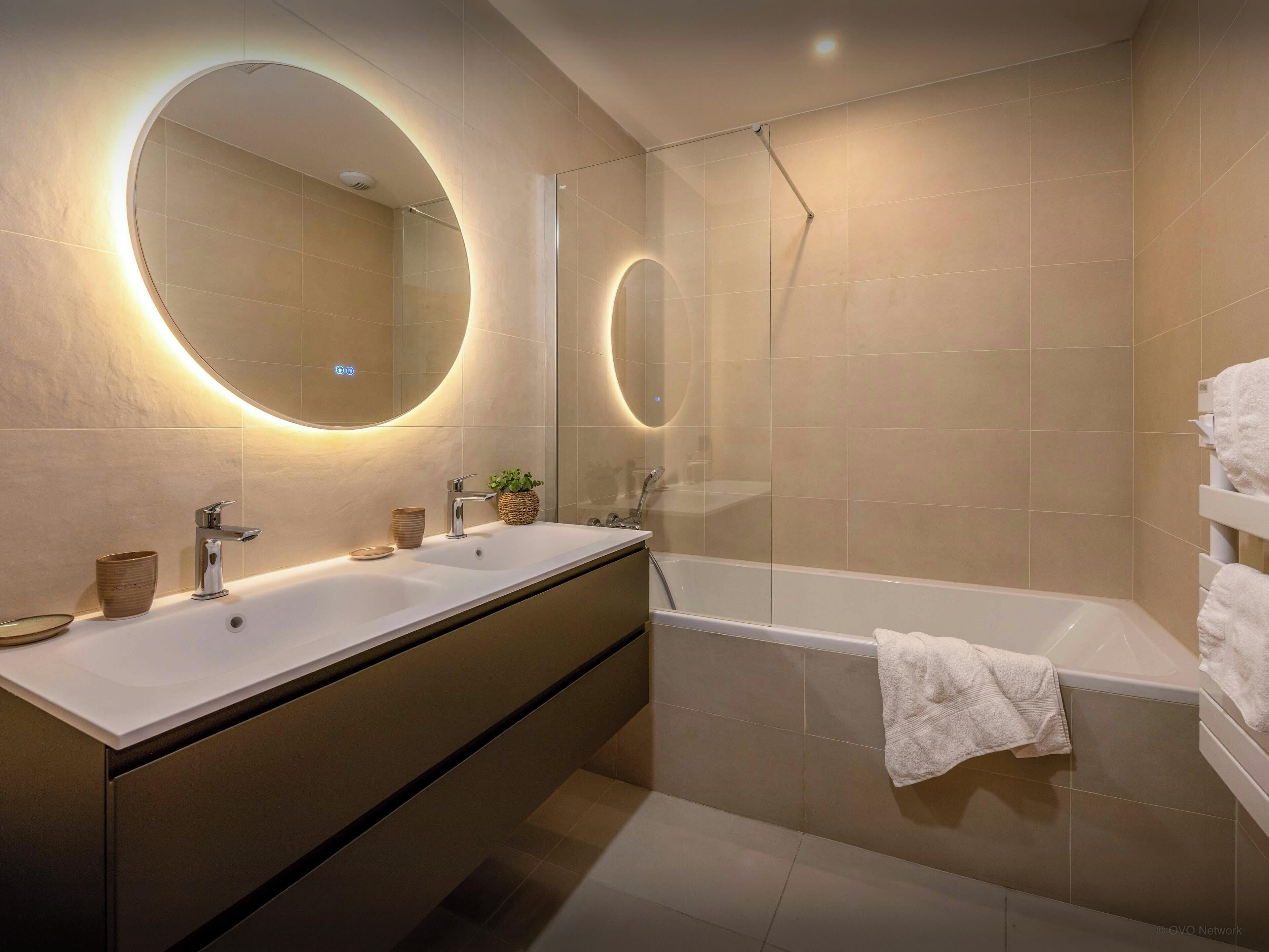 Un baño moderno con tocador doble, bañera y un espejo grande con iluminación integrada.