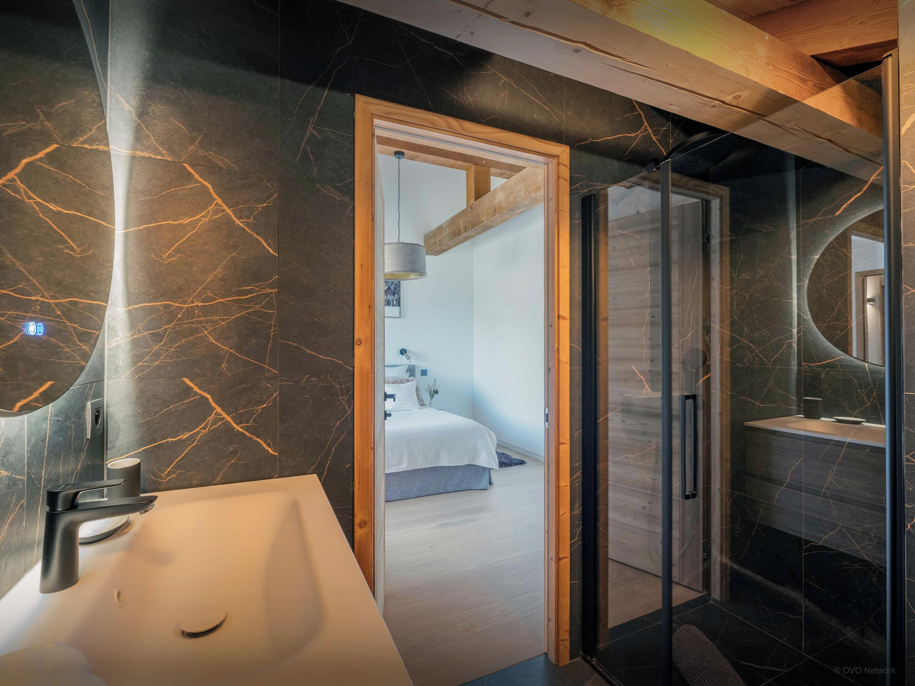 Une salle de bain moderne avec un meuble-lavabo en marbre, une cabine de douche en verre et une chambre visible, équipée d’un lit et d’une table de chevet.