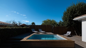 Ein rechteckiger Swimmingpool mit Holzterrasse, zwei Liegestühlen und einem Holenzaun.