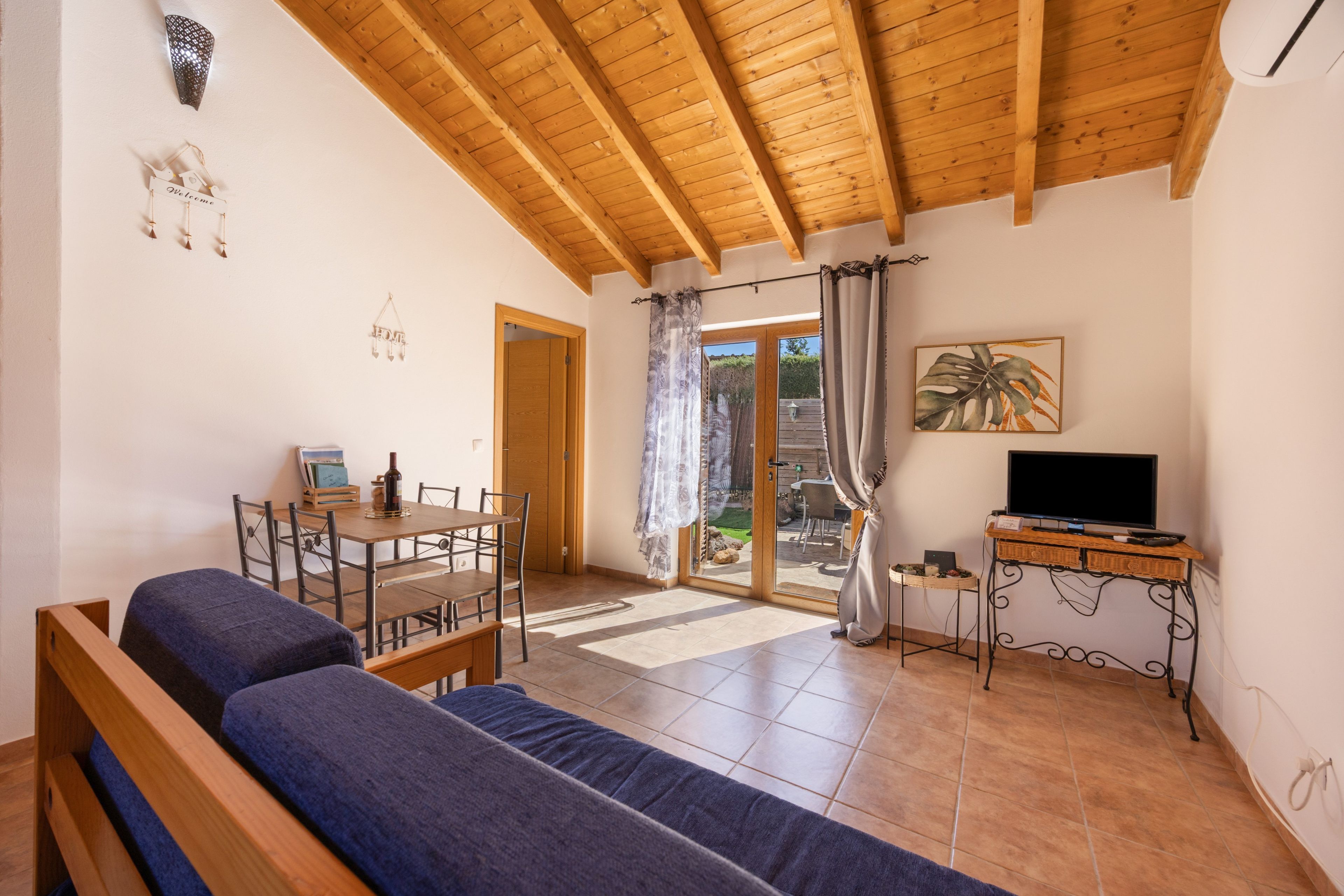 A cozy room with a wooden ceiling, a sofa, a dining table with chairs, a TV on a stand, and a door leading outside.