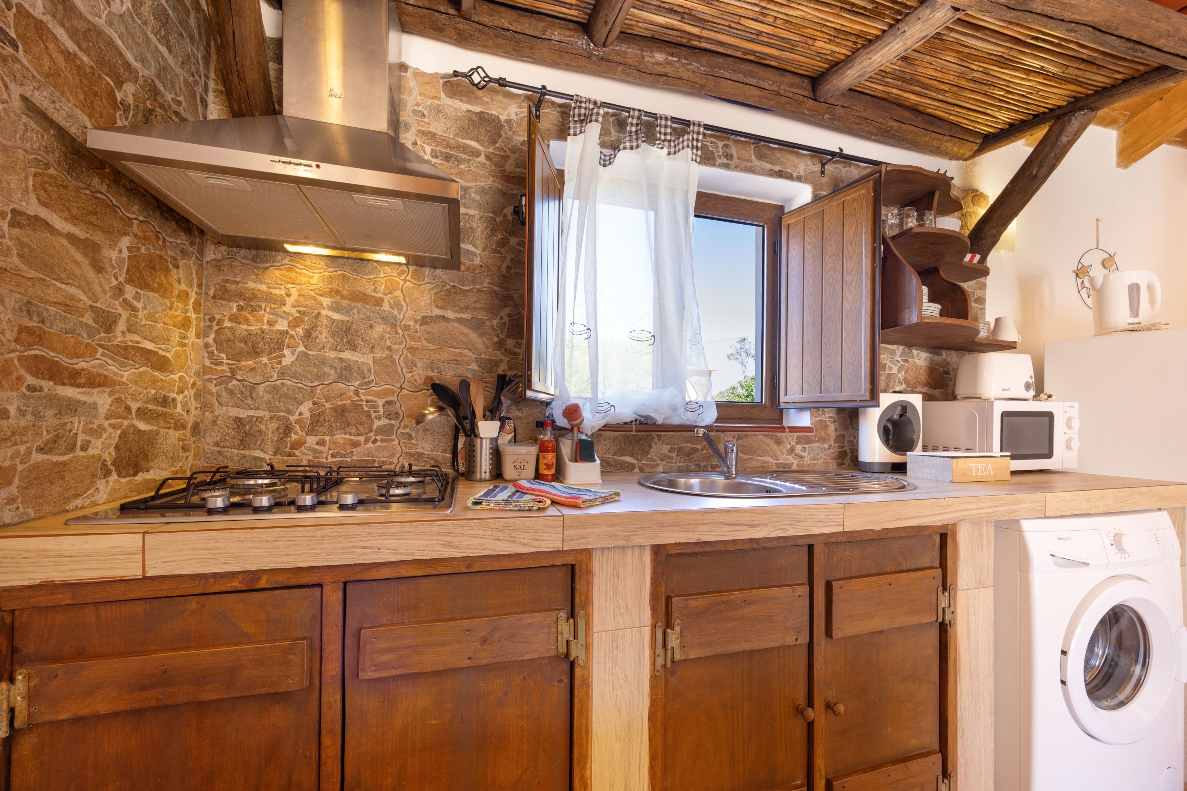 A rustic kitchen with a stone wall, wooden cabinets, a gas stove, a sink, and a washing machine.