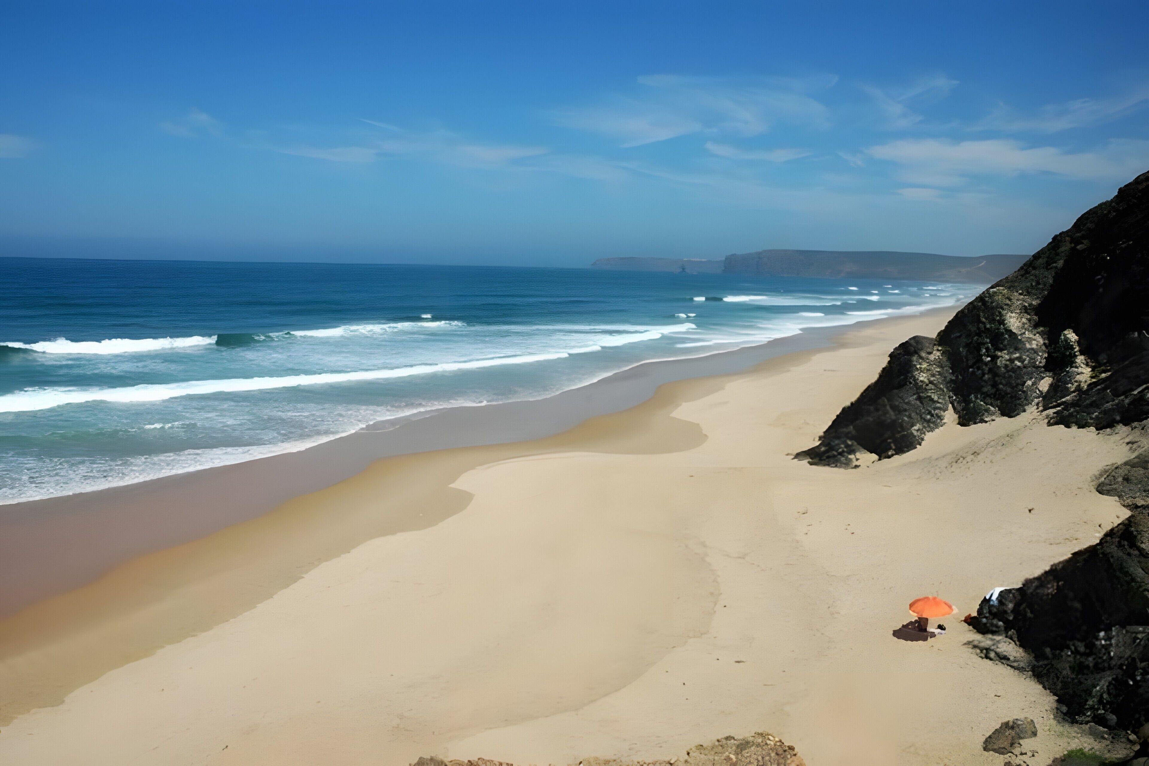 Uma praia com céu claro, ondas e um penhasco rochoso.