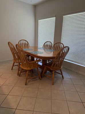 A dining area with a round wooden table surrounded by six chairs, a tiled floor, and two windows with blinds. - The Lake Breeze Villa- 48GH (Laguna Vista)