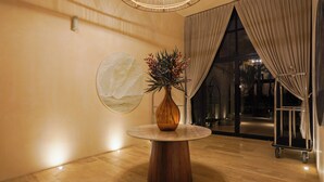 A round wooden table with a vase of flowers, a wall-mounted light fixture, and a view of the outdoors through a large window with curtains.